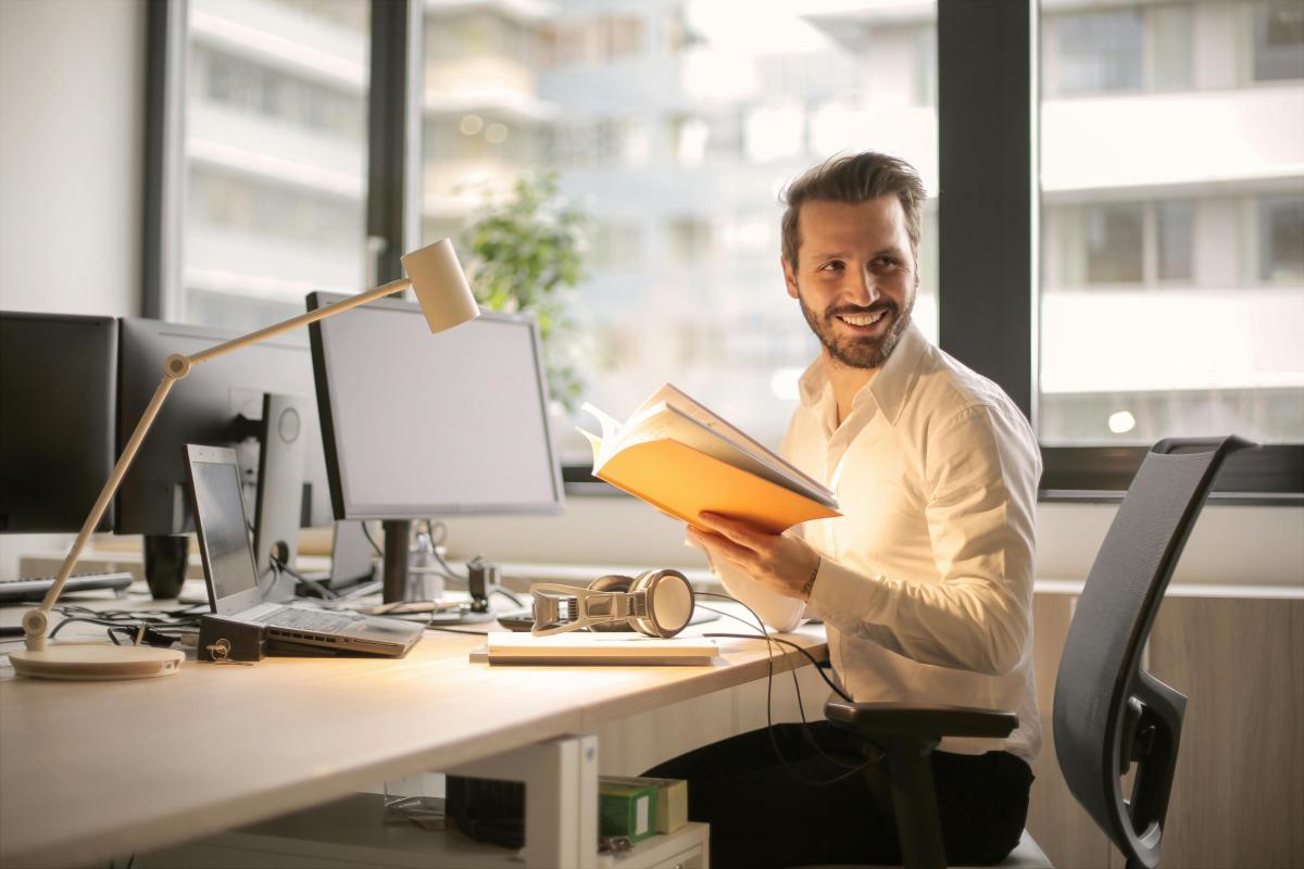 Laechelnder Mann am Schreibtisch mit Handbuch aks Symbolbild fuer das Training: Hybrid Cloud Fundamentals (NHCF) v7.5 Kurs Online